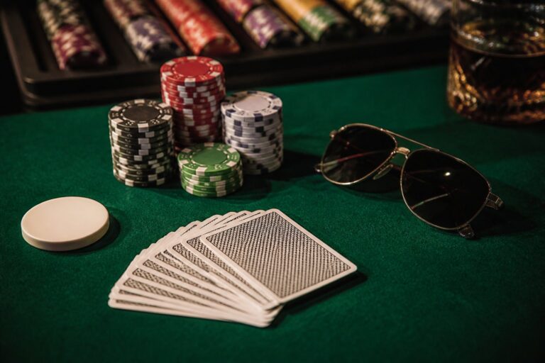 Poker chips, a stack of playing cards face down, a white dealer button, sunglasses, and a glass of drink on a green felt table.
